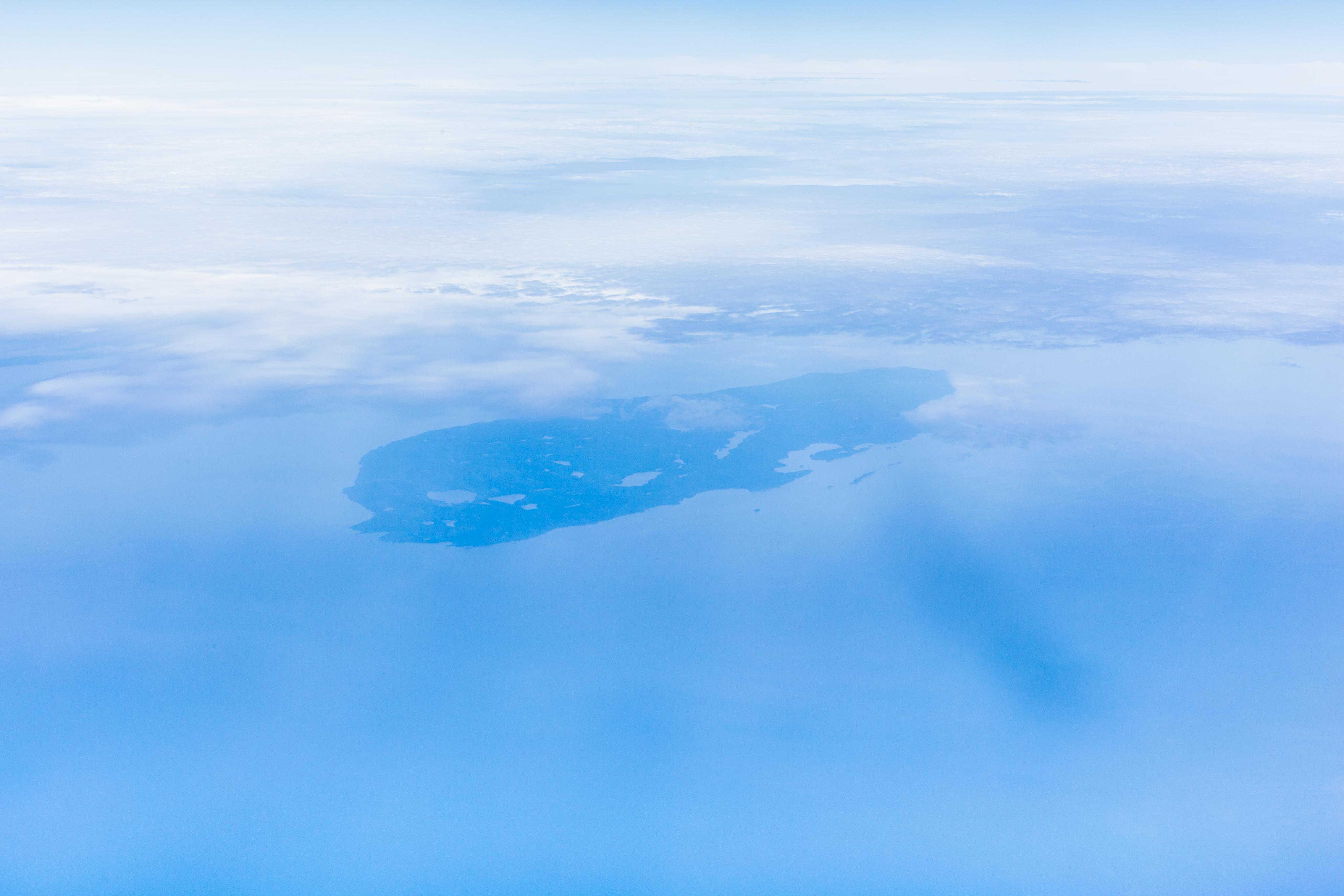A very low contrast, light blue aerial image of the entire island of Isle Royale. From a perspective of roughly 30,000 feet. Clouds obscure parts of Lake Superior.
